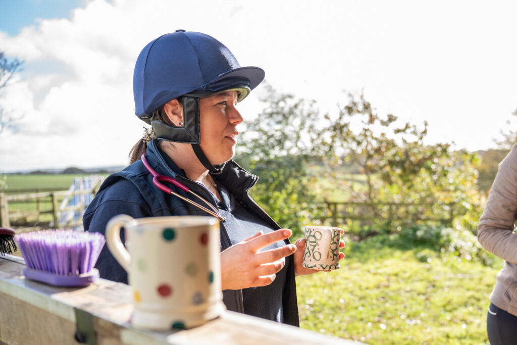 A person wearing a riding helmet and a stethoscope holds a mug outdoors, standing beside a table with mugs and a purple brush. Sunlit greenery and a fence are visible in the background.