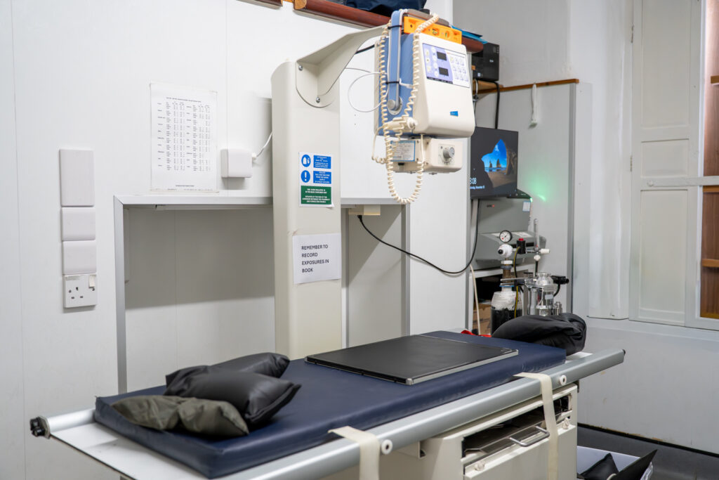 A modern X-ray machine in a medical examination room, featuring a padded table, control panel, and various medical equipment on the walls and nearby surfaces.