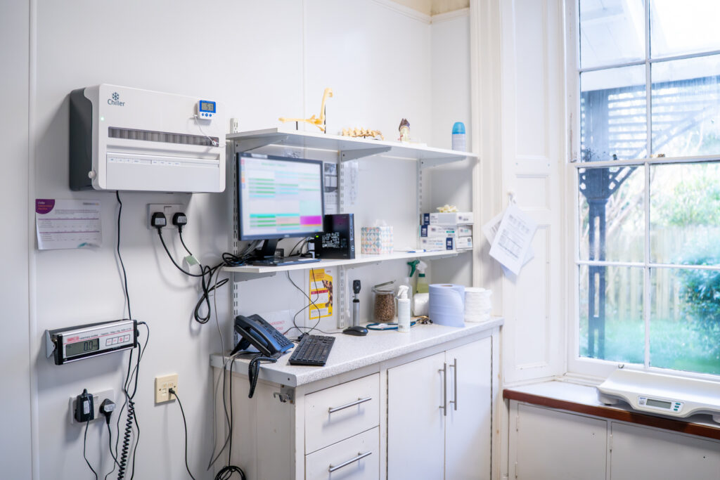 A tidy medical surgery with a computer, telephone, shelves with supplies, and a window letting in natural light. Cupboards are below the worktop, and various medical equipment hangs on the wall. A garden is visible outside.