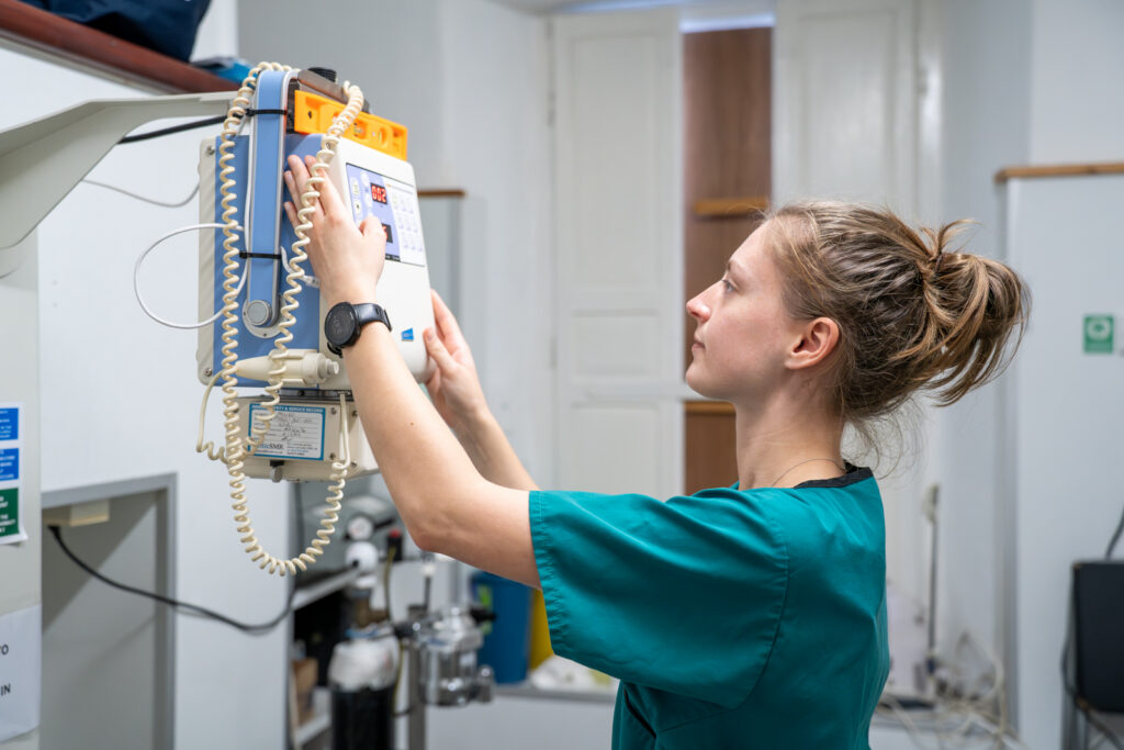 A healthcare worker in teal scrubs adjusts a medical device mounted on a wall in a clinical setting. She is standing sideways, focused on the equipment, with her hair tied back.