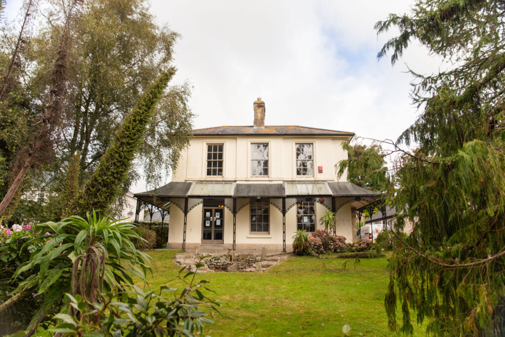 A two-storey cream-coloured house with large windows, a metal roof covering a wide veranda, and surrounded by green trees and a well-kept lawn.