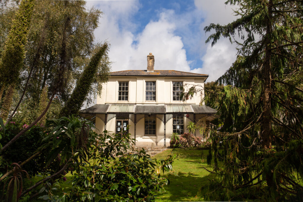 A cream-coloured two-storey house with tall windows and a covered porch, surrounded by lush green trees and bushes under a partly cloudy sky.