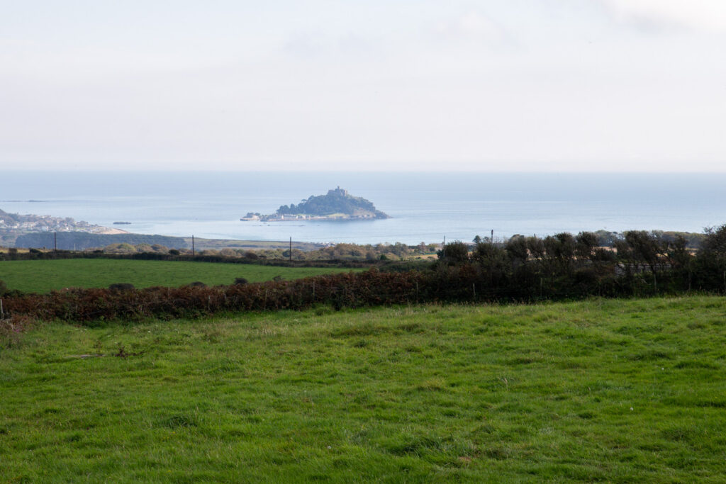 A distant view of an island with a castle on it, surrounded by the sea, seen from a grassy field with hedges and a cloudy sky above.