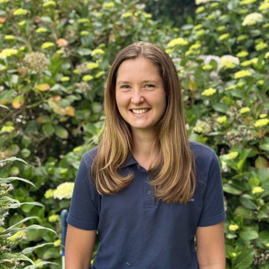 Meet the team: A young woman with long, straight brown hair, wearing a navy blue polo shirt, sits and smiles in front of lush green bushes with blooming hydrangeas.