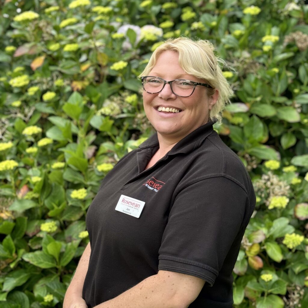Meet the team: A smiling woman with blonde hair and glasses stands outdoors in front of green leafy plants and yellow flowers, wearing a black polo shirt and a name badge that reads Roseann.