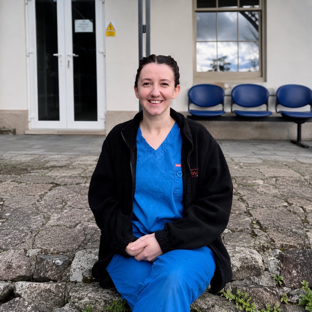 Meet the team: A woman in blue medical scrubs and a black jacket sits on stone steps outside a building, smiling at the camera. Empty blue chairs and a glass door are visible in the background.