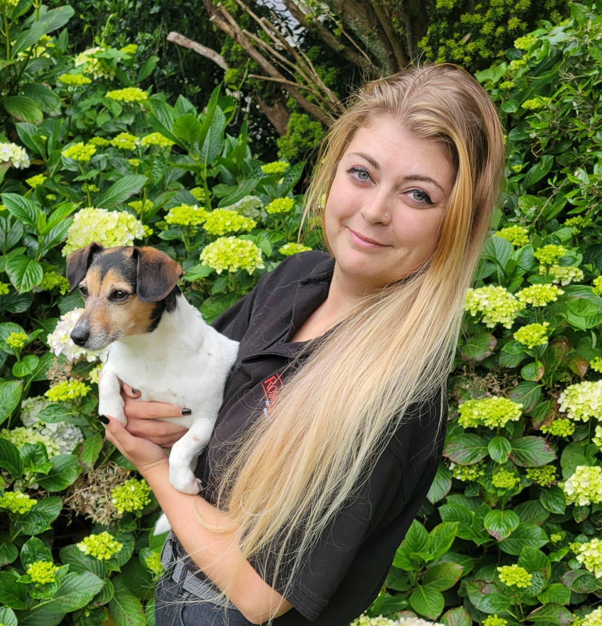 Meet the team: A woman with long blonde hair holds a small black, brown, and white dog. They are standing in front of green bushes with light green and white flowers.