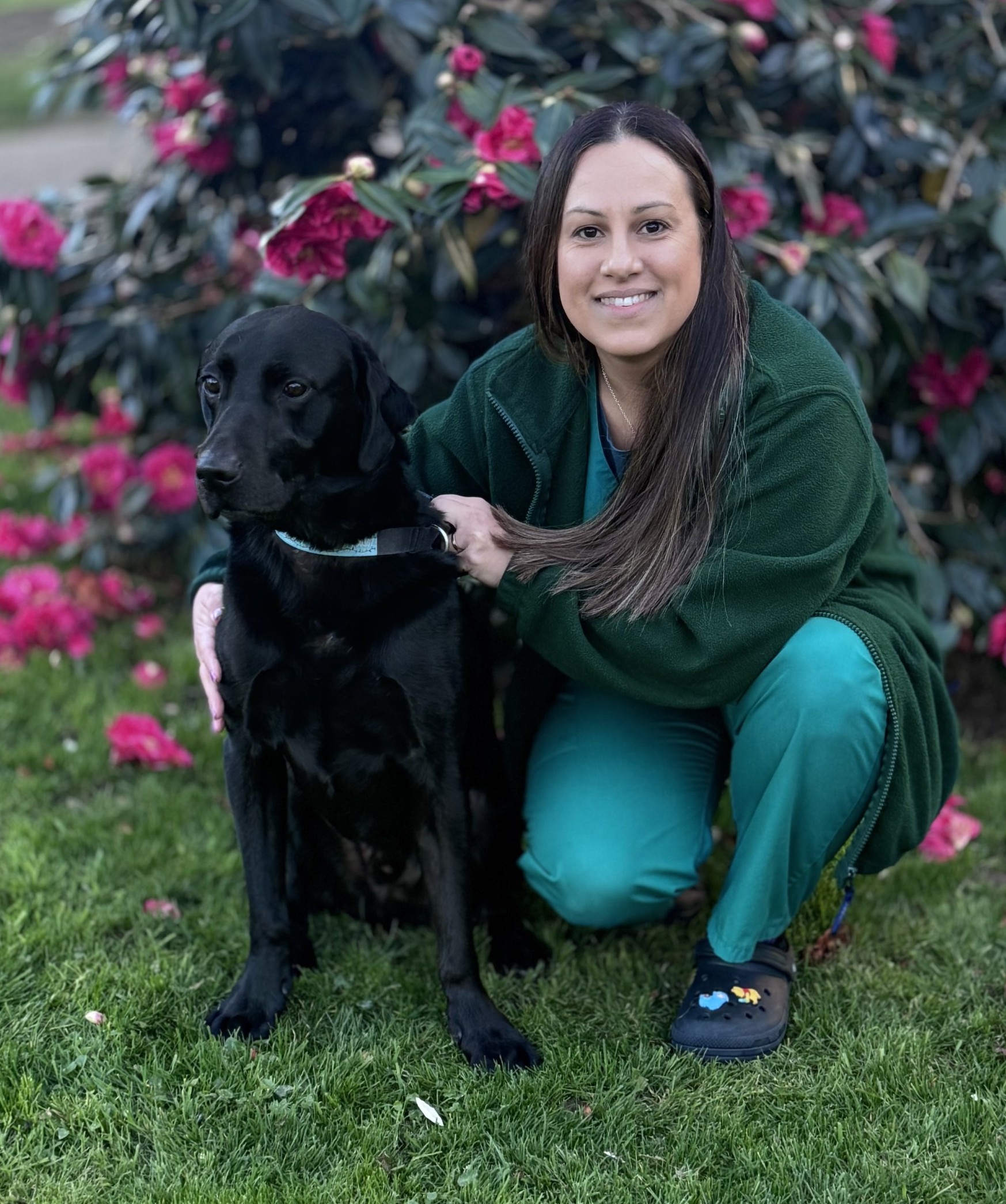 Meet the team: A woman in green scrubs kneels on grass next to a large black dog, holding its collar. They are outdoors in front of a bush with pink flowers.