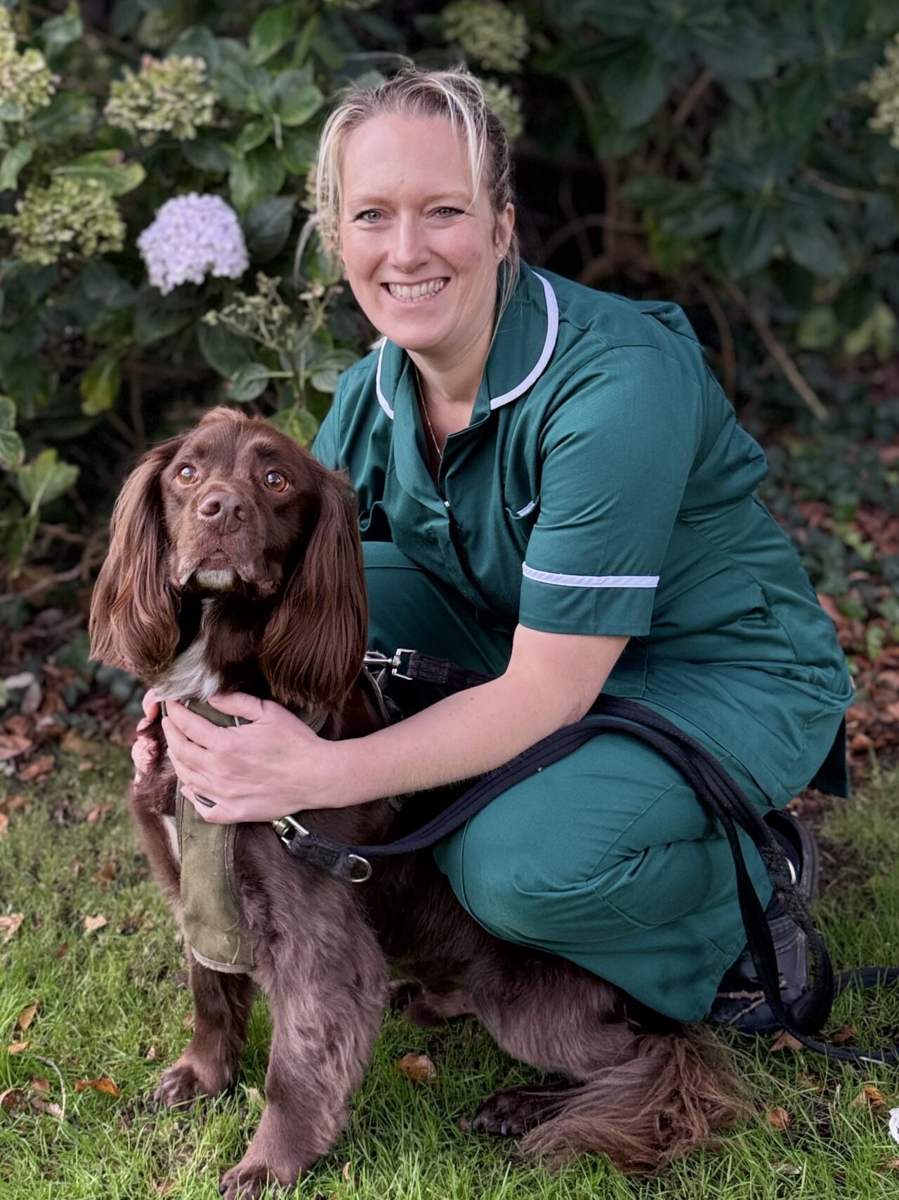 Meet the team: A woman in a green uniform kneels on grass, smiling and holding a brown dog on a lead. They are outdoors in front of leafy bushes with light-coloured flowers.