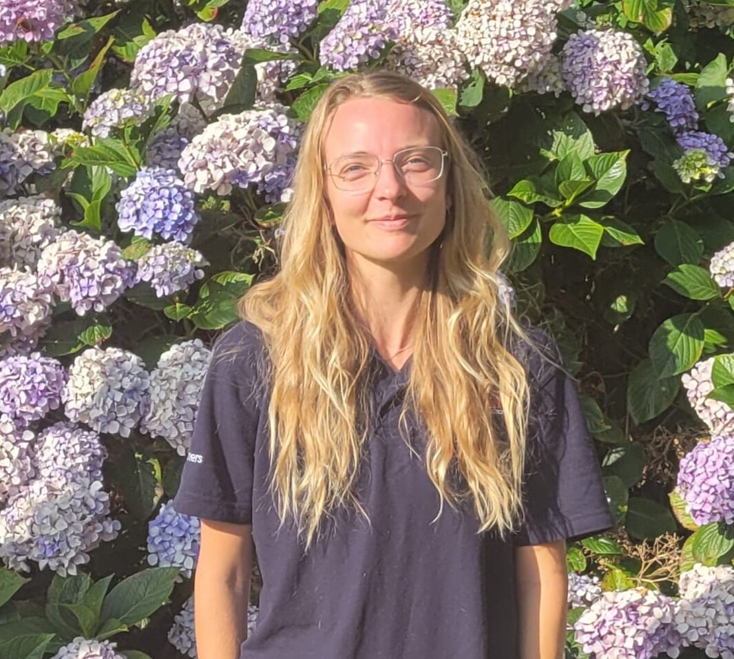 Meet the team: A person with long blonde hair and glasses stands in front of a lush wall of blooming purple and white hydrangeas, wearing a dark shirt and light shorts, hands in pockets, smiling in the sunlight.